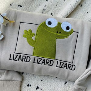 Puede incluir: Sudadera beige con un diseño de lagarto verde dentro de un rectángulo negro. El texto "LIZARD LIZARD LIZARD" está bordado debajo. La sudadera tiene puños acanalados y cuello redondo.