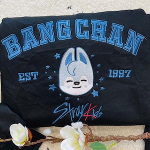 May include: Black sweatshirt with blue embroidered text and a cartoon character. The text reads "BANG CHAN" above the character, with "EST 1997" below. The words "Stray Kids" are embroidered in blue below the character.
