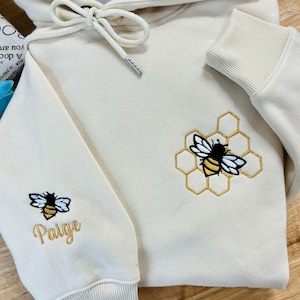 Puede incluir: Una sudadera con capucha de color crema con un diseño de abeja y panal bordado en la manga derecha. La manga izquierda tiene una abeja y el nombre "Paige" bordado.