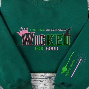 May include: A dark green sweatshirt with the text "YOU WILL BE CHANGED WICKED FOR GOOD". The sweatshirt has a pink crown, a witch hat, a green broom, and a pink wand with stars embroidered on it.