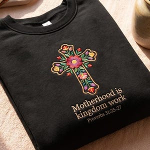 May include: Black sweatshirt featuring an embroidered floral cross design. The cross is adorned with colorful flowers and leaves. The text "Motherhood is kingdom work Proverbs 31:25-27" is printed below the cross.