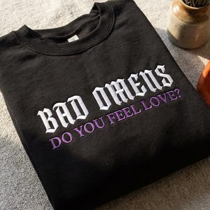 Puede incluir: Sudadera negra de cuello redondo con el texto bordado "BAD OMENS DO YOU FEEL LOVE?" en blanco y morado. Una bufanda decorativa y un pequeño jarrón con lavanda están en el fondo.