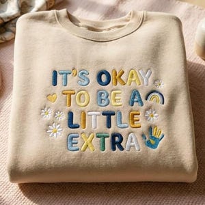 Op de afbeelding: Beige sweatshirt met de geborduurde tekst "IT'S OKAY TO BE A LITTLE EXTRA" in blauw, geel en groen. Het ontwerp bevat een regenboog, bloemen en een handgebaar. Een sjaal en een vaas met bloemen staan op de achtergrond.