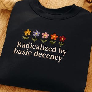 Op de afbeelding: Marineblauwe sweatshirt met geborduurde bloemen in geel, paars, roze, oranje en rood. De tekst "Radicalized by basic decency" is eronder geborduurd. De sweatshirt is gemaakt van zacht materiaal.
