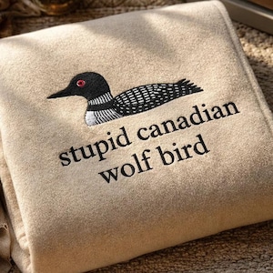 Pode incluir: Um cobertor bege dobrado com um desenho bordado de um pássaro preto e branco com um olho vermelho. O texto "stupid canadian wolf bird" é bordado abaixo.