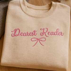 May include: A beige sweatshirt featuring the pink embroidered words "Dearest Reader" and a bow. The folded sweatshirt rests on a wooden surface. A camera, headphones, and a mug are also visible in the image.