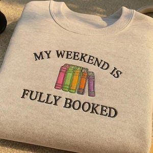May include: A pale grey sweatshirt featuring the embroidered phrase "MY WEEKEND IS FULLY BOOKED" above a row of colourful books. The books are in shades of pink, green, yellow, orange, and purple. The sweatshirt has a crew neck.