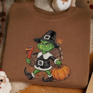 May include: Brown sweatshirt with an embroidered Thanksgiving-themed design. The design features a green character in a pilgrim hat holding a turkey and a pumpkin. The sweatshirt is surrounded by holiday decorations.