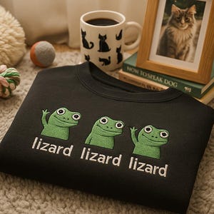 Könnte beinhalten: Schwarzes Sweatshirt mit drei grünen Eidechsen-Cartoon-Grafiken und dem Wort "lizard" darunter. Ein gerahmtes Katzenfoto, eine Tasse mit Katzensilhouetten und ein Hundespielzeug befinden sich im Hintergrund.