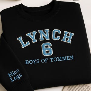 Peut inclure: Sweat-shirt noir avec broderie "LYNCH 6 BOYS OF TOMMEN" bleu clair. La manche porte la broderie "Nice Legs" en bleu clair. Une tasse à café blanche et des bobines de fil sont en arrière-plan.