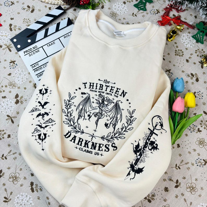 May include: A cream-colored sweatshirt with a black graphic print of a winged creature and the text "the THIRTEEN FROM NOW UNTIL THE DARKNESS +CLAIMS US+"