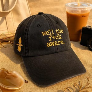 May include: Black baseball cap with the embroidered text "well the f*ck aware" in yellow. A gold dragon graphic is on the side. The cap is on a sandy surface.