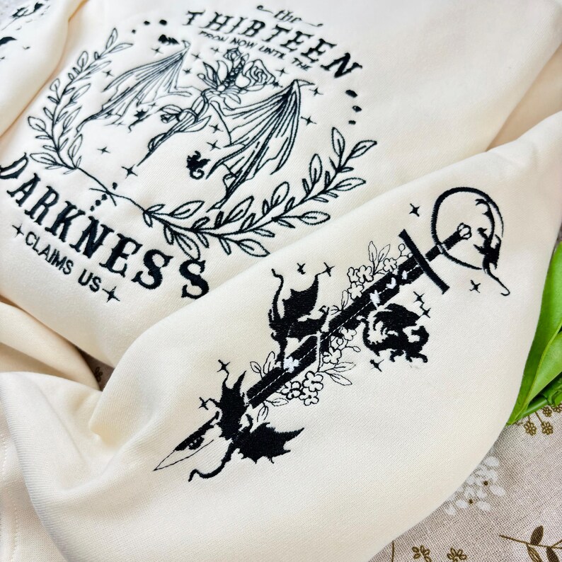 May include: A white sweatshirt with black embroidered designs. The front features a dragon with the text "the thirteen from now until the... darkness claims us". The sleeve features a sword with a dragon and floral designs.