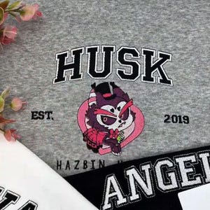 Pode incluir: Um moletom cinza com a palavra "HUSK" em letras pretas e brancas acima de um personagem de desenho animado. O personagem est&aacute; dentro de um cora&ccedil;&atilde;o rosa. O texto "EST. 2019" e "HAZBIN" tamb&eacute;m s&atilde;o vis&iacute;veis.