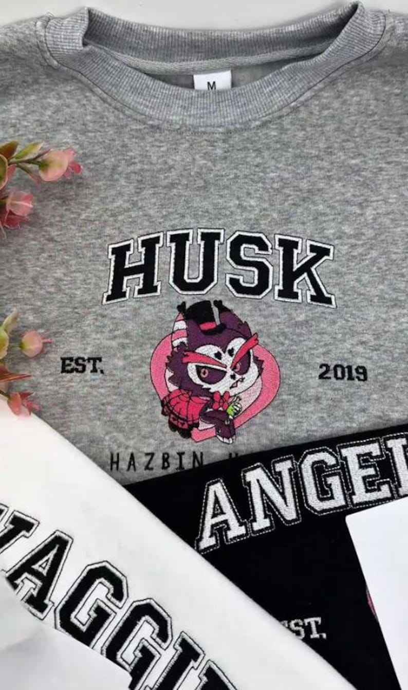 Pode incluir: Um moletom cinza com a palavra "HUSK" em letras pretas e brancas acima de um personagem de desenho animado. O personagem est&aacute; dentro de um cora&ccedil;&atilde;o rosa. O texto "EST. 2019" e "HAZBIN" tamb&eacute;m s&atilde;o vis&iacute;veis.