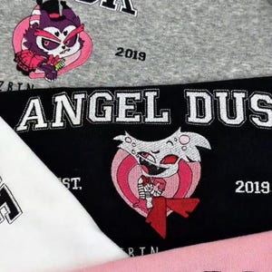 Pode incluir: Close-up de camisolas bordadas em cinza, preto, branco e rosa. As camisolas apresentam as palavras "Husk", "Angel Dust" e "Charlie" em uma fonte universit&aacute;ria, juntamente com desenhos de personagens de desenhos animados e o ano "2019".