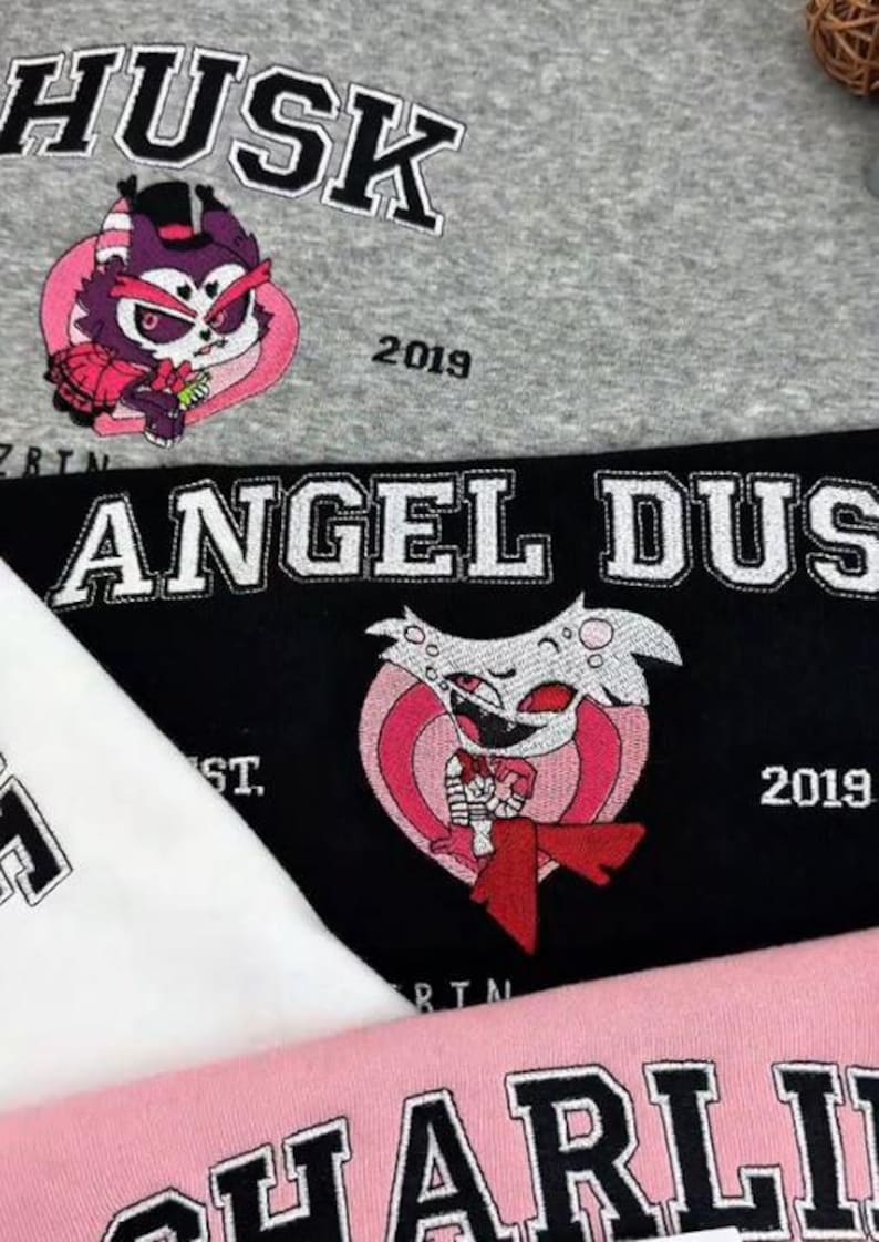 Pode incluir: Close-up de camisolas bordadas em cinza, preto, branco e rosa. As camisolas apresentam as palavras "Husk", "Angel Dust" e "Charlie" em uma fonte universit&aacute;ria, juntamente com desenhos de personagens de desenhos animados e o ano "2019".
