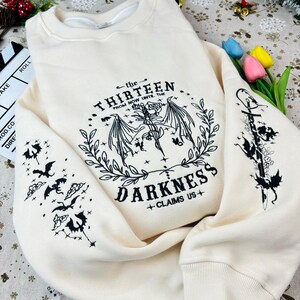 May include: A cream-colored sweatshirt with black embroidered designs. The front features a winged creature with the text "the THIRTEEN from NOW UNTIL THE DARKNESS +CLAIMS US+". The sleeves feature smaller winged creatures.