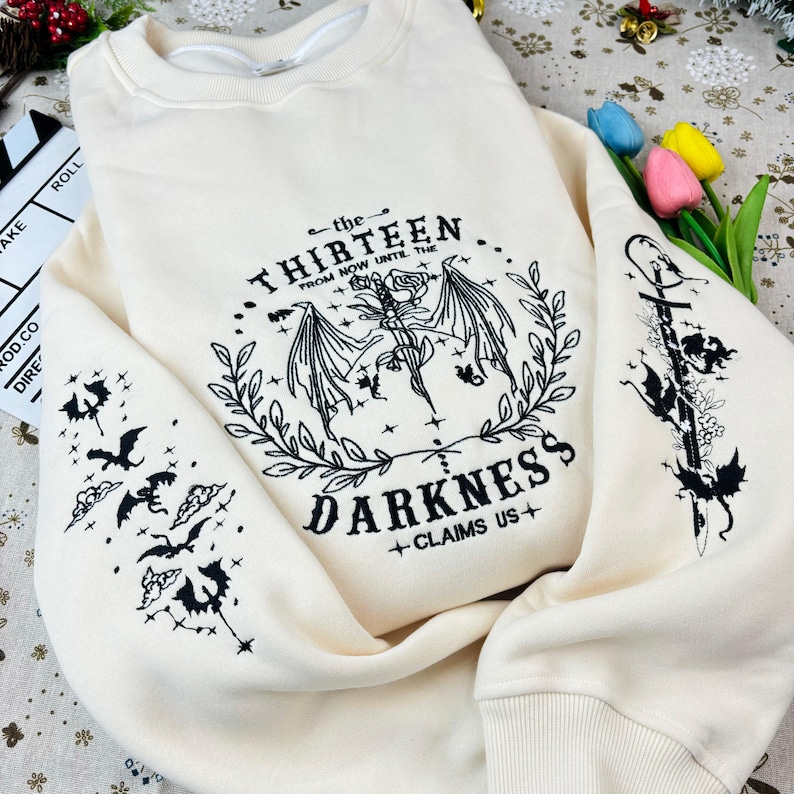May include: A cream-colored sweatshirt with black embroidered designs. The front features a winged creature with the text "the THIRTEEN from NOW UNTIL THE DARKNESS +CLAIMS US+". The sleeves feature smaller winged creatures.