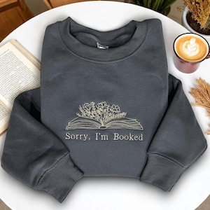Puede incluir: Una sudadera gris oscuro con el texto bordado "Sorry, I'm Booked" sobre un diseño de libro abierto con detalles florales. La sudadera está doblada sobre una superficie blanca, con un libro y una taza de café en el fondo.