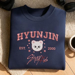 May include: Navy blue sweatshirt with the name "HYUNJIN" in pink embroidered lettering. Below the name is a white cat face with pink accents, surrounded by pink stars. The text "EST. 2000" and "Stray Kids" are also embroidered in pink.
