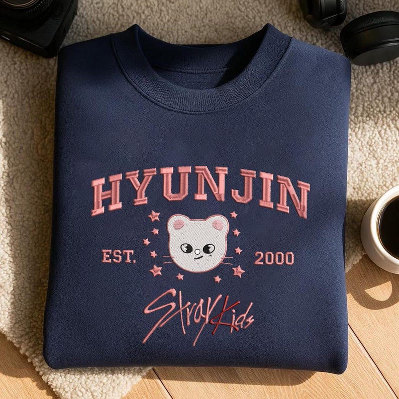 May include: Navy blue sweatshirt with the name "HYUNJIN" in pink embroidered lettering. Below the name is a white cat face with pink accents, surrounded by pink stars. The text "EST. 2000" and "Stray Kids" are also embroidered in pink.