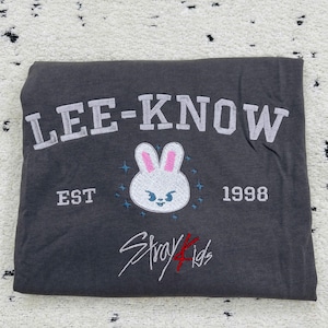 May include: Dark gray t-shirt with the text "LEE-KNOW" in white block letters. Below is a white bunny face with pink ears and blue accents, the text "EST 1998", and the words "Stray Kids" in red and white script.
