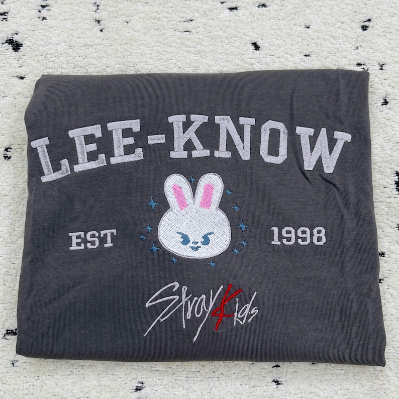 May include: Dark gray t-shirt with the text "LEE-KNOW" in white block letters. Below is a white bunny face with pink ears and blue accents, the text "EST 1998", and the words "Stray Kids" in red and white script.