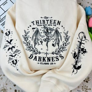May include: A cream-colored sweatshirt with black embroidered designs. The front features a winged creature with a sword and a rose, surrounded by a wreath. The text "the THIRTEEN FROM NOW UNTIL THE DARKNESS +CLAIMS US+" is also embroidered on the front. The sleeves have embroidered designs of winged creatures and swords.
