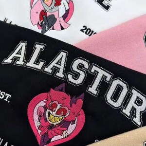 Pode incluir: Close-up de tecido bordado com personagens de desenhos animados e texto. A palavra "VAGGIE" &eacute; costurada em letras pretas sobre um fundo branco, com um personagem de desenho animado. A palavra "ALASTOR" &eacute; costurada em um fundo preto.