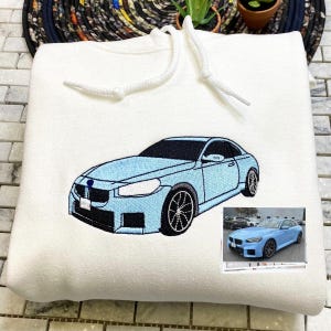 May include: A white hoodie with a blue embroidered car on the front. The car is a sedan with a black interior and a black grill. The hoodie has a white drawstring.
