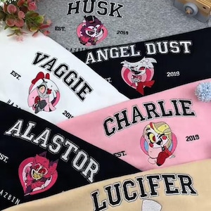 Pode incluir: Uma cole&ccedil;&atilde;o de camisolas de gola redonda em v&aacute;rias cores, cada uma bordada com o nome de um personagem e imagem de Hazbin Hotel. As camisolas apresentam os nomes HUSK, VAGGIE, ANGEL DUST, CHARLIE, ALASTOR e LUCIFER, com o ano de 2019.