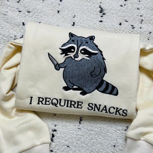 May include: Cream-colored sweatshirt featuring an embroidered raccoon holding a knife. The raccoon is gray and black with a striped tail. The text "I REQUIRE SNACKS" is below the raccoon in black lettering.