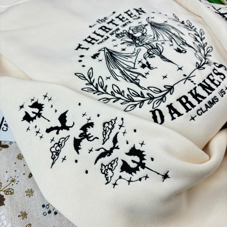 May include: A white sweatshirt with black embroidered designs. The front of the sweatshirt features a large design with the text "the THIRTEEN from now until the... DARKNESS + CLAIMS US". The sleeve has a smaller design with bats and clouds.