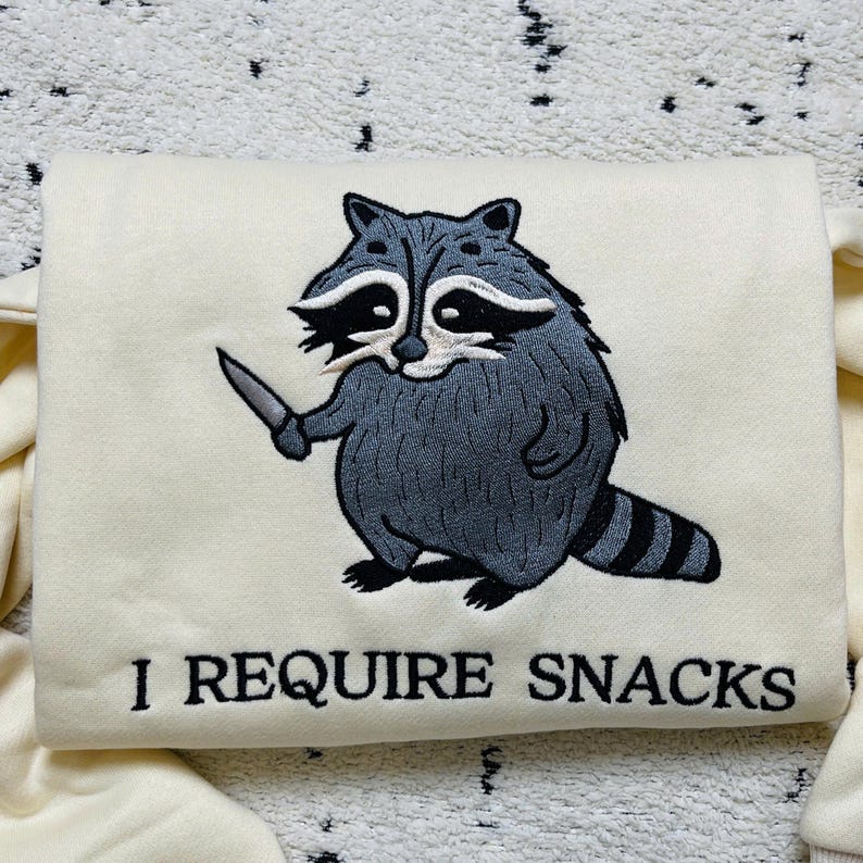 May include: Cream-colored sweatshirt featuring an embroidered raccoon holding a knife. The raccoon is gray and white with a black striped tail. The text "I REQUIRE SNACKS" is below the raccoon.