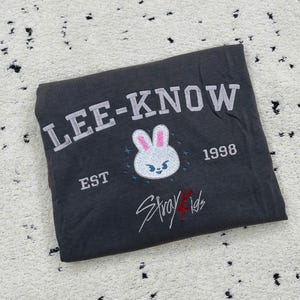 May include: Dark gray t-shirt with the text "LEE-KNOW" in white block letters. Below is a white bunny face with pink ears and blue accents, the text "EST 1998", and the words "Stray Kids" in red script.