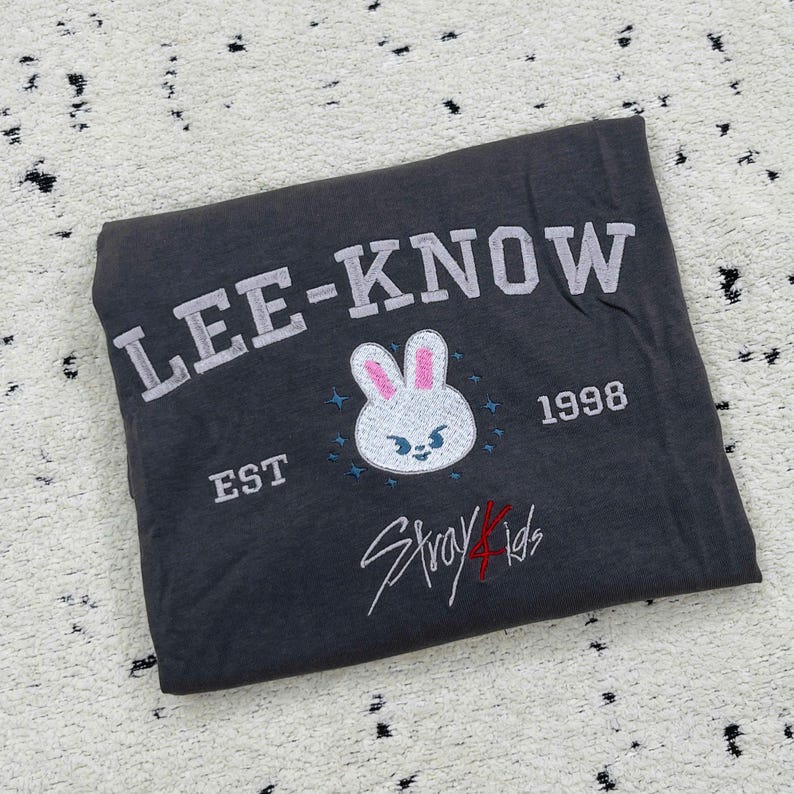 May include: Dark gray t-shirt with the text "LEE-KNOW" in white block letters. Below is a white bunny face with pink ears and blue accents, the text "EST 1998", and the words "Stray Kids" in red script.