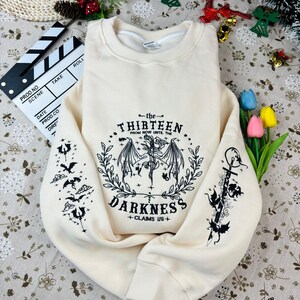 May include: A cream-colored sweatshirt with a black graphic print of a winged creature and the text "the THIRTEEN from NOW UNTIL THE DARKNESS +CLAIMS US+"