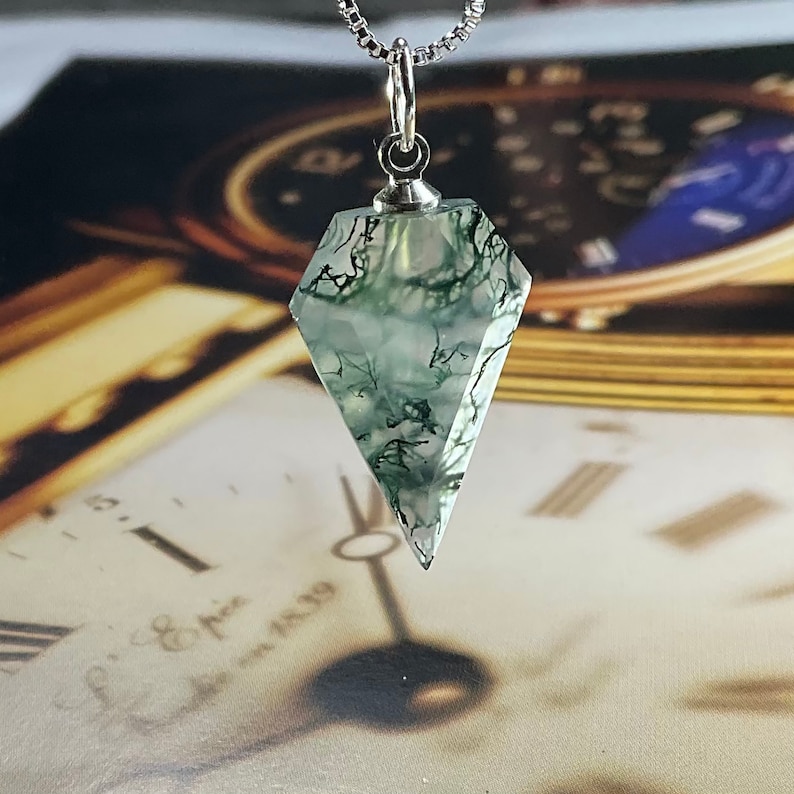 May include: A silver chain necklace with a green moss agate pendant in the shape of a diamond. The pendant is clear with green moss-like inclusions.