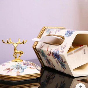 May include: A white ceramic tissue box cover with a floral design and gold accents. The cover has a deer figurine on top and a slot for pulling out tissues.