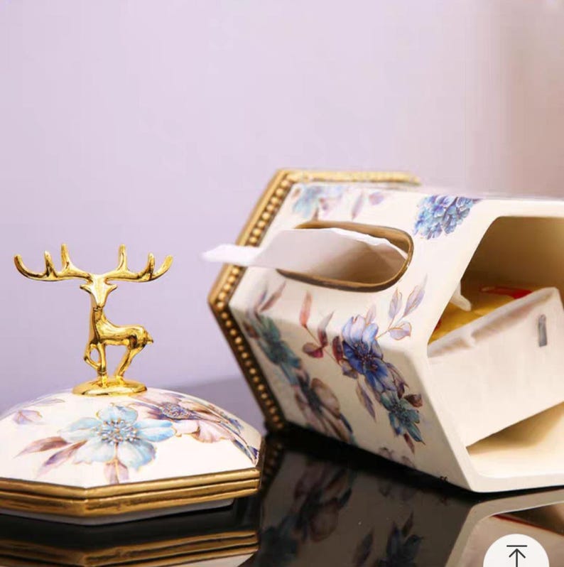 May include: A white ceramic tissue box cover with a floral design and gold accents. The cover has a deer figurine on top and a slot for pulling out tissues.