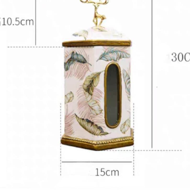 May include: White ceramic tissue box cover with a gold trim and a deer figurine on top. The box has a floral pattern with pink and green leaves. The box measures 10.5 inches tall, 15 inches wide, and 30 inches deep.