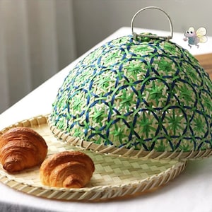 May include: A woven bamboo food cover with a green and blue pattern. The cover has a handle and is sitting on a woven bamboo tray with two croissants.