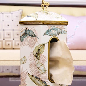 May include: A white ceramic tissue box cover with a gold trim and a gold deer figurine on top. The cover has a tropical leaf pattern and a slot for tissues.