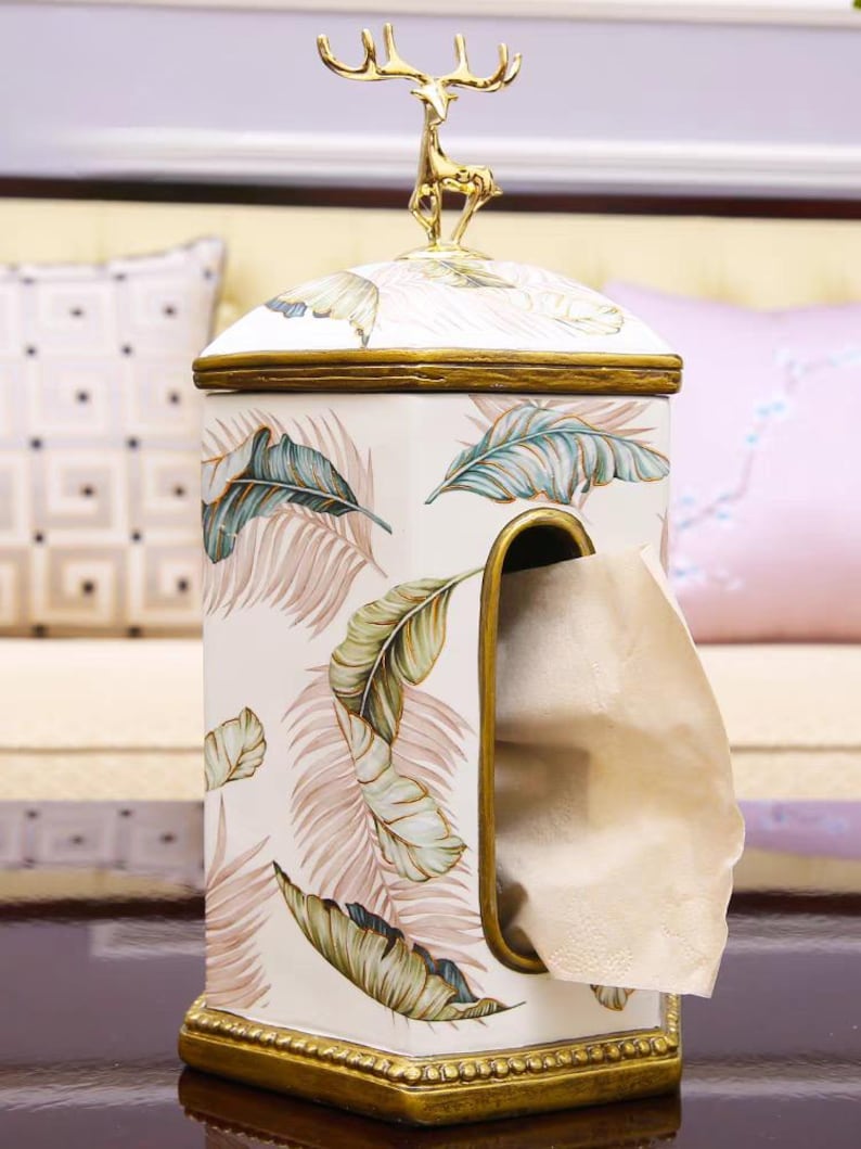 May include: A white ceramic tissue box cover with a gold trim and a gold deer figurine on top. The cover has a tropical leaf pattern and a slot for tissues.