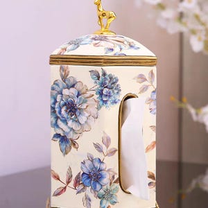 May include: White ceramic tissue box cover with a gold deer figurine on top. The box is decorated with blue and white floral designs.