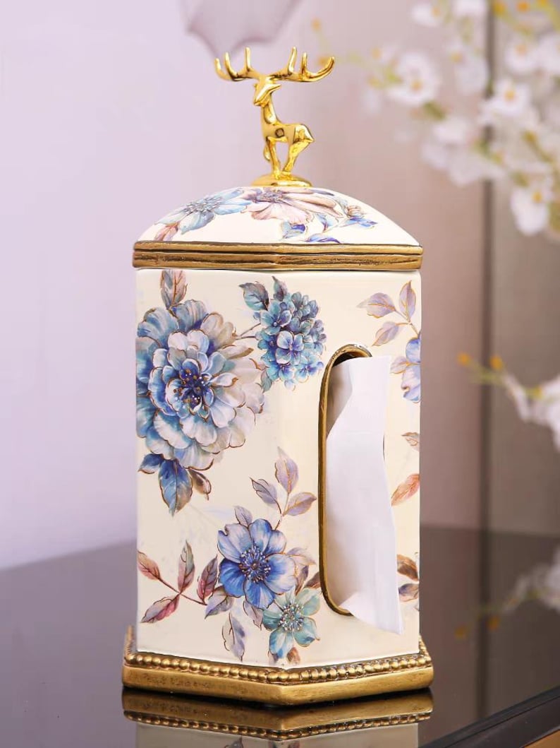 May include: White ceramic tissue box cover with a gold deer figurine on top. The box is decorated with blue and white floral designs.