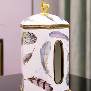 May include: White ceramic tissue box cover with a gold deer figurine on top. The box has a gold trim and a blue and white feather pattern.