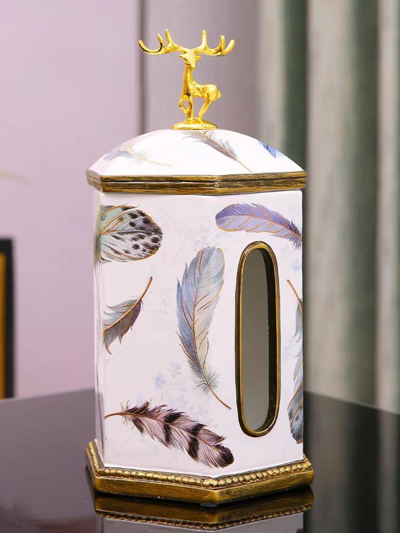 May include: White ceramic tissue box cover with a gold deer figurine on top. The box has a gold trim and a blue and white feather pattern.