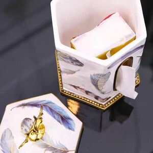 May include: White ceramic tissue box holder with a hexagonal shape and a gold trim. The holder has a lid with a gold knob and a feather design. The holder is filled with white tissues.
